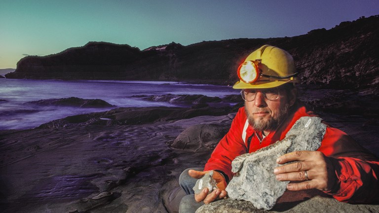 A man crouching down on a beach, wearing a hard hat, holding two rocky fossils